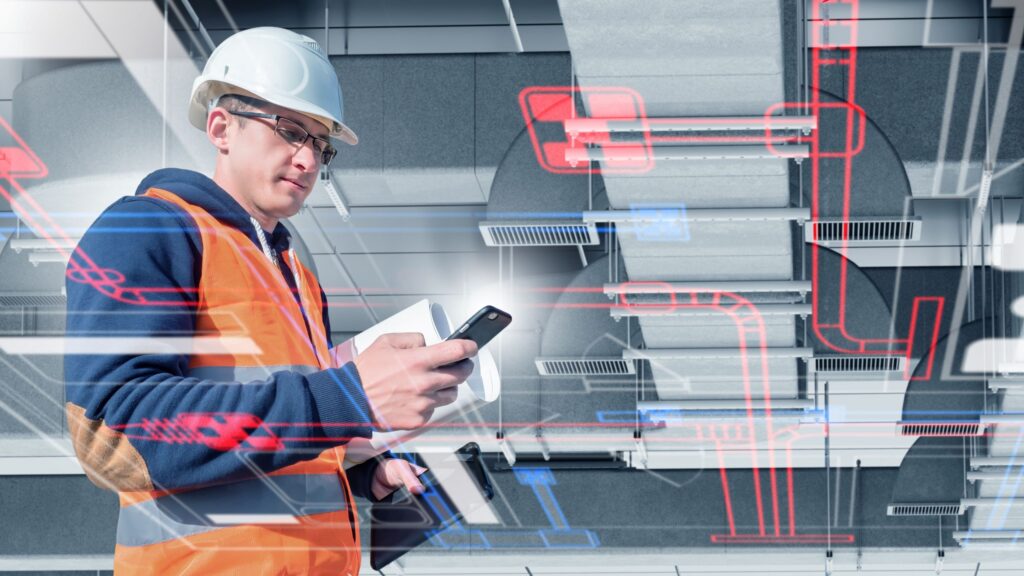 Bauleiter mit Schutzhelm und Warnweste hält Smartphone und Tablet vor einer Decke mit technischen Rohrleitungen und digitalen Überlagerungen